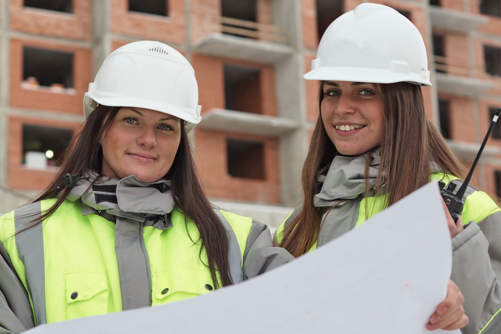 Zwei Bauingenieurinnen mit Schutzhelmen und Bauplänen auf einer Baustelle